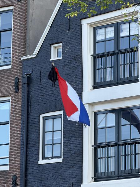 A striking dark blue Amsterdam canal house stands prominently on Staalkade, Centrum, Amsterdam, adorned with a Dutch flag flying beneath a black bag, suggesting a moment of national remembrance